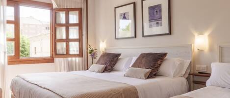 A double bed with a beige bedspread, a nightstand with a lamp, and a window with curtains.