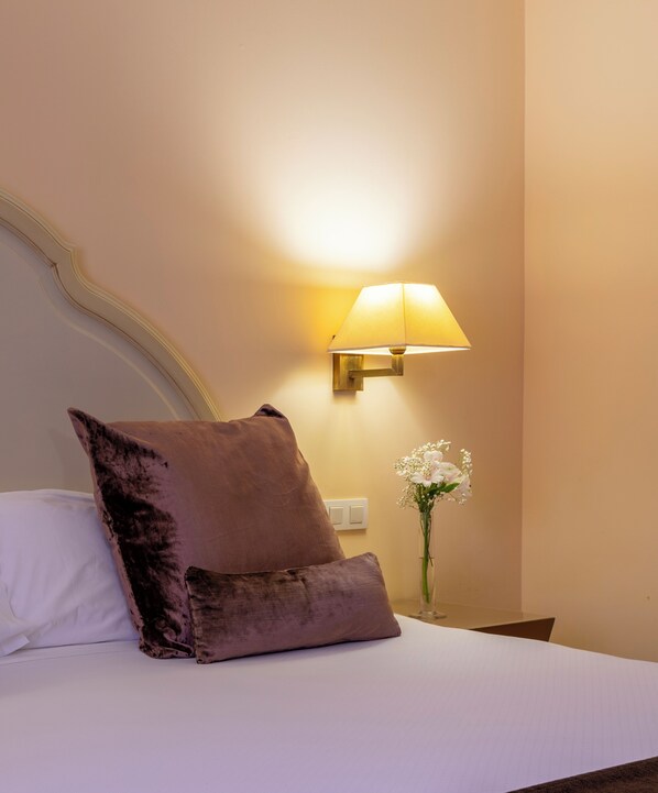A neatly made bed with a large purple pillow and a smaller one beside it. A wall-mounted lamp provides light, and a vase with white flowers is on the nightstand.