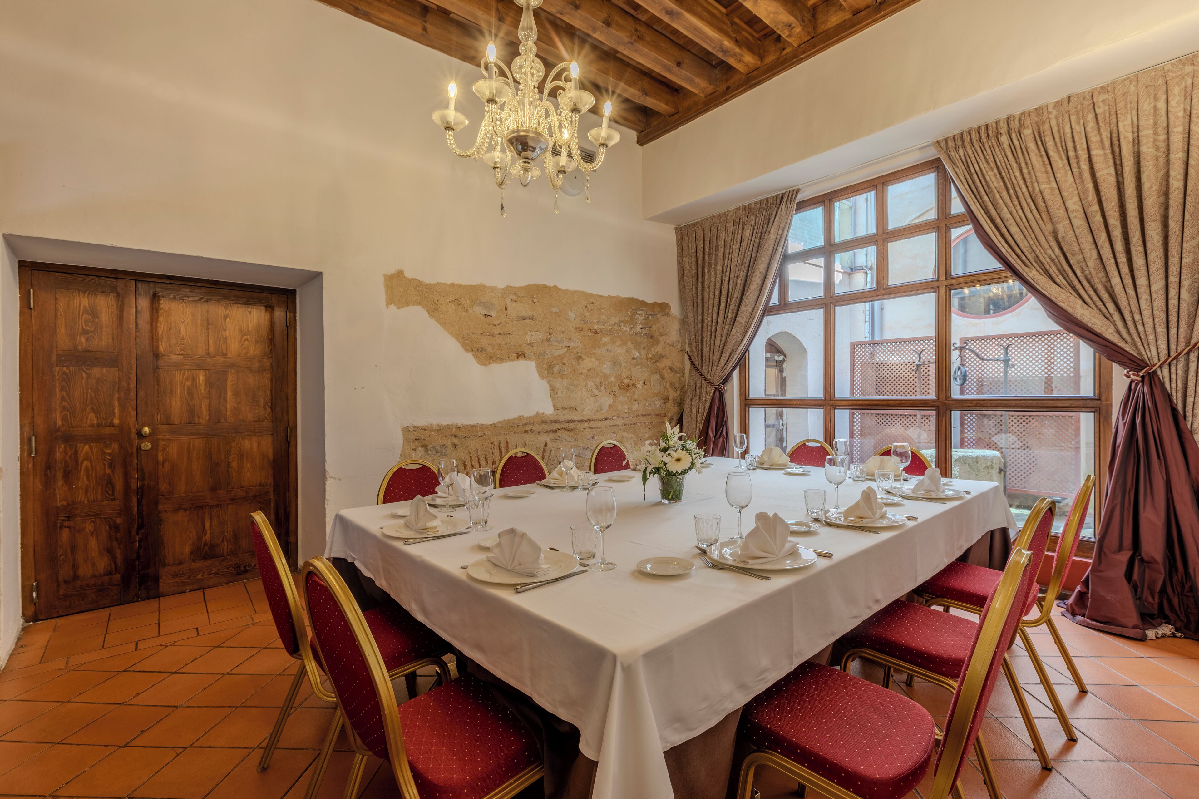 A dining area with a large table set for a meal, red upholstered chairs, a chandelier, and a view through a window with curtains.