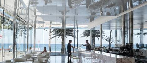Un bar moderno con mostrador de mármol, asientos de estructura metálica y grandes ventanales con vistas al mar.