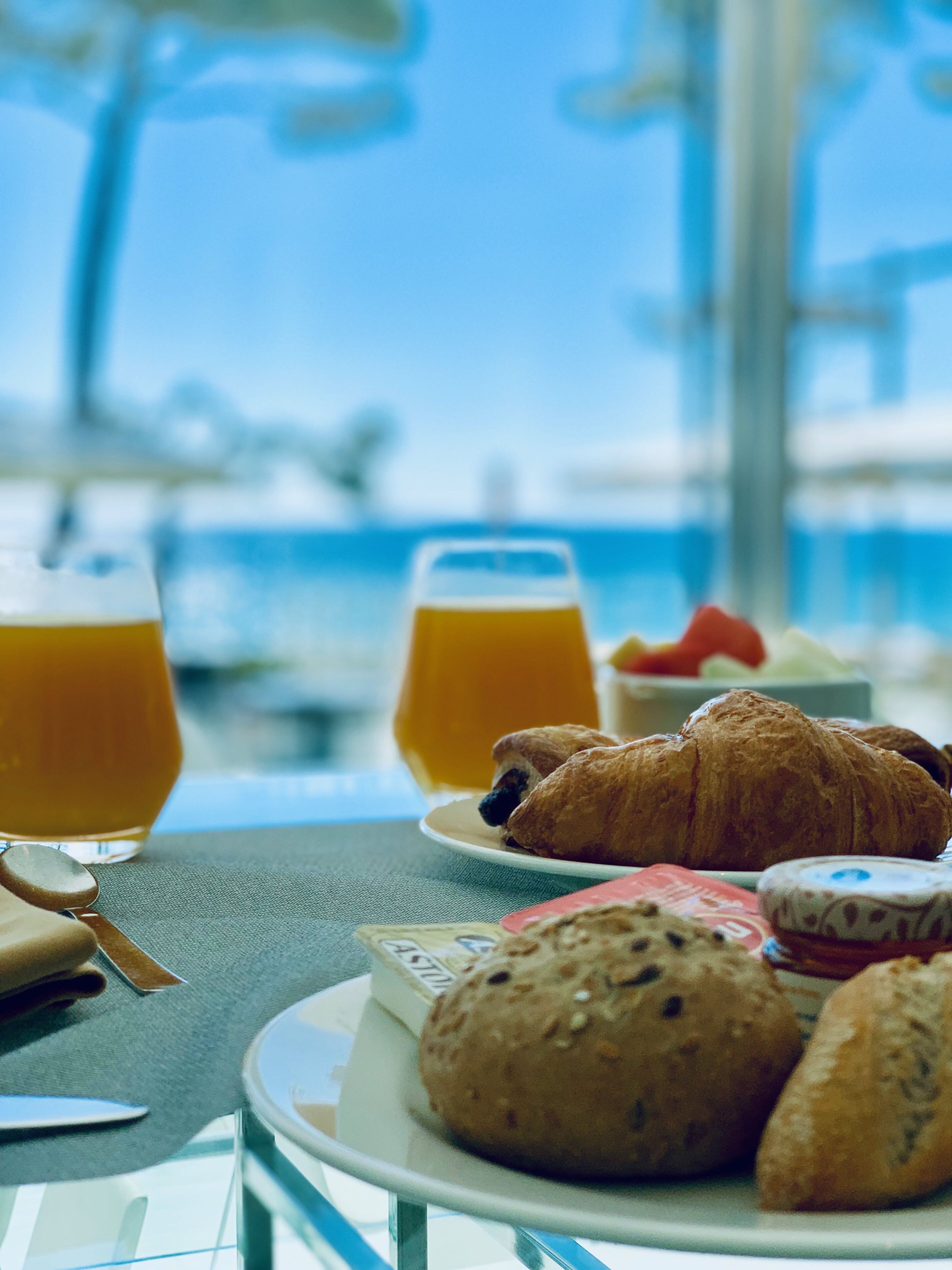 Frühstückstisch mit Croissants, Muffins und Orangensaft, mit Blick auf das Meer und Schiffe im Hintergrund.