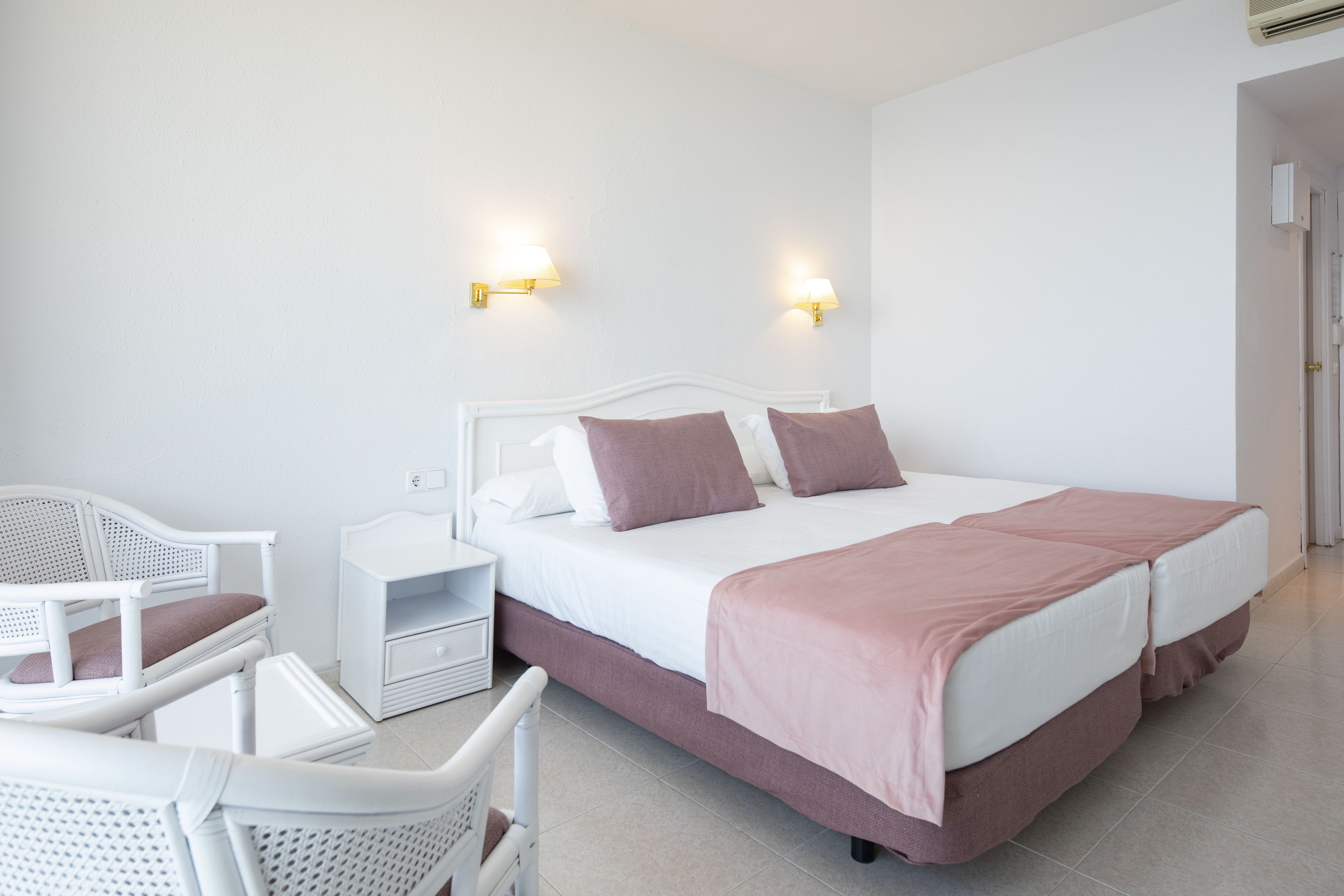 A hotel room with a white bed, two bedside tables, a white chair, and two wall-mounted lamps.