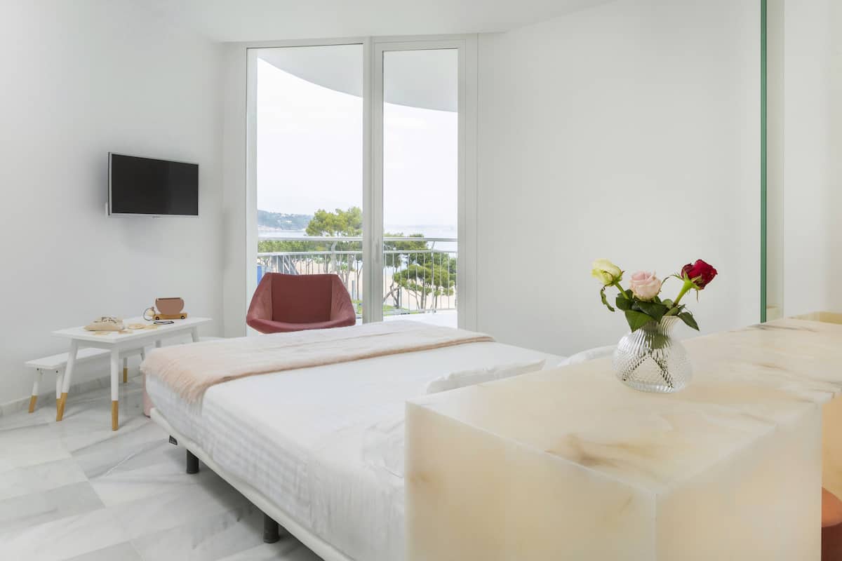 Ein modernes Hotelzimmer mit einem großen Bett, einem kleinen Schreibtisch, einem Stuhl, einem an der Wand montierten Fernseher und einem Balkon mit Blick.
