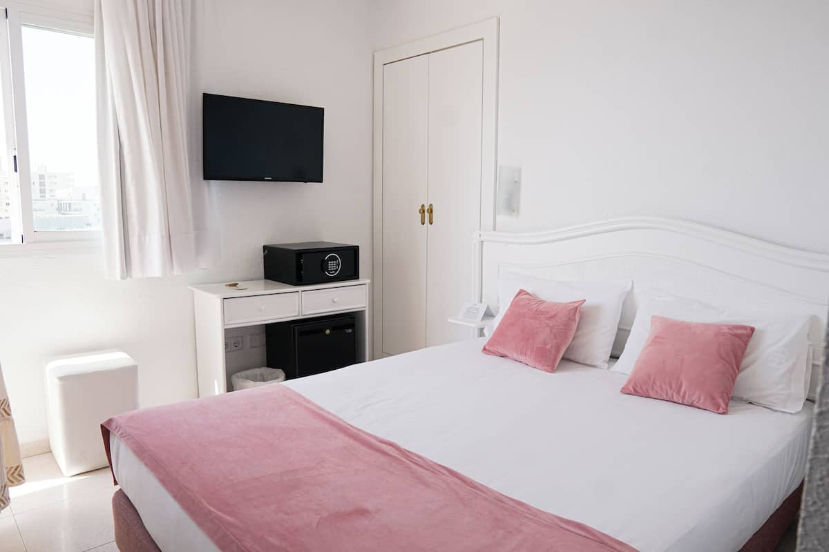 Ein Hotelzimmer mit einem Bett, einem an der Wand befestigten Fernseher, einem Schreibtisch mit Computer und einem Fenster mit Vorhängen.