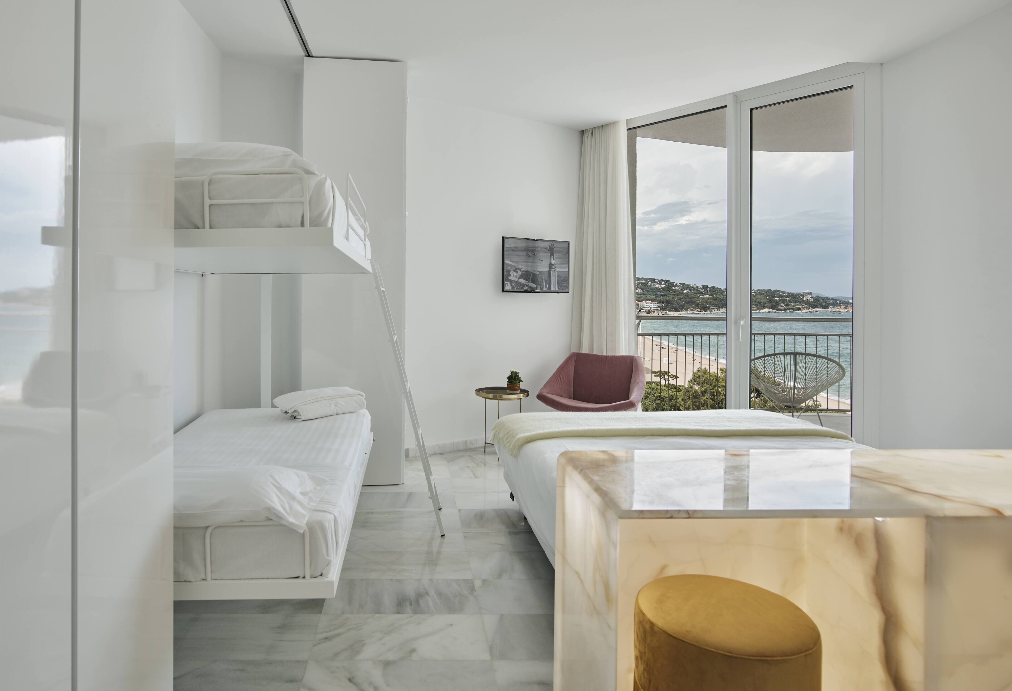Ein modernes Hotelzimmer mit Etagenbett, Einzelbett, Holztisch, rotem Stuhl und großem Fenster mit Blick auf Meer und Hügel.