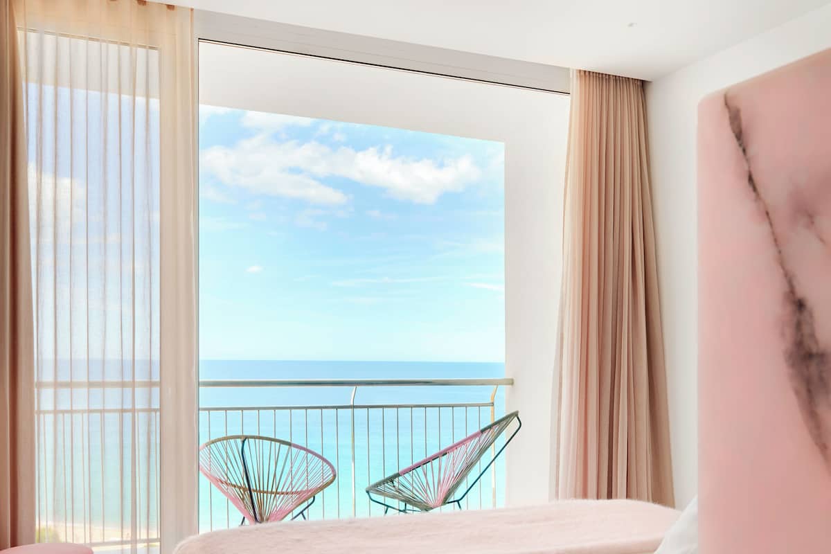 Ein Schlafzimmer mit großem Fenster, einem Balkon und einem Bett mit einem pinken Pouf.