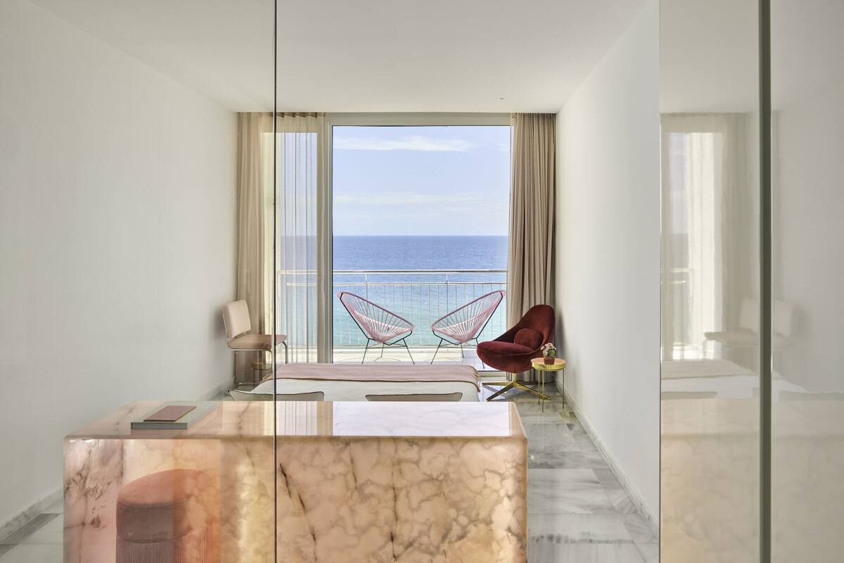 Ein modernes Hotelzimmer mit einem Marmor-Schreibtisch, zwei Stühlen und einem großen Fenster mit Meerblick.