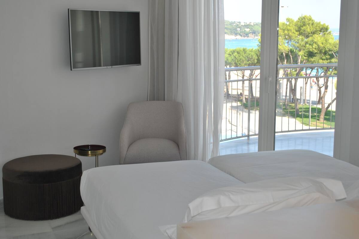 Ein Hotelzimmer mit Bett, Sessel, rundem Hocker, Fernseher und einem Balkon mit Blick auf das Meer und Bäume.