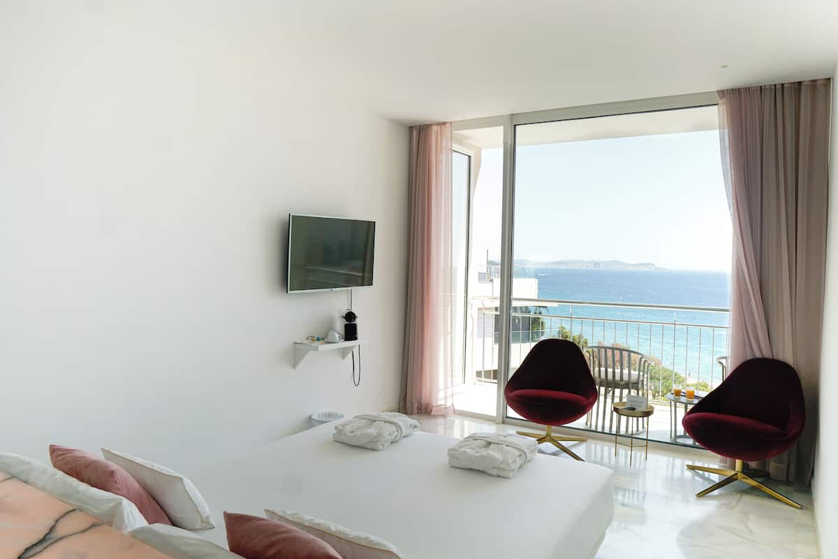 Ein Hotelzimmer mit einem großen Bett, zwei roten Sesseln, einem Fernseher und einem Balkon mit Meerblick.