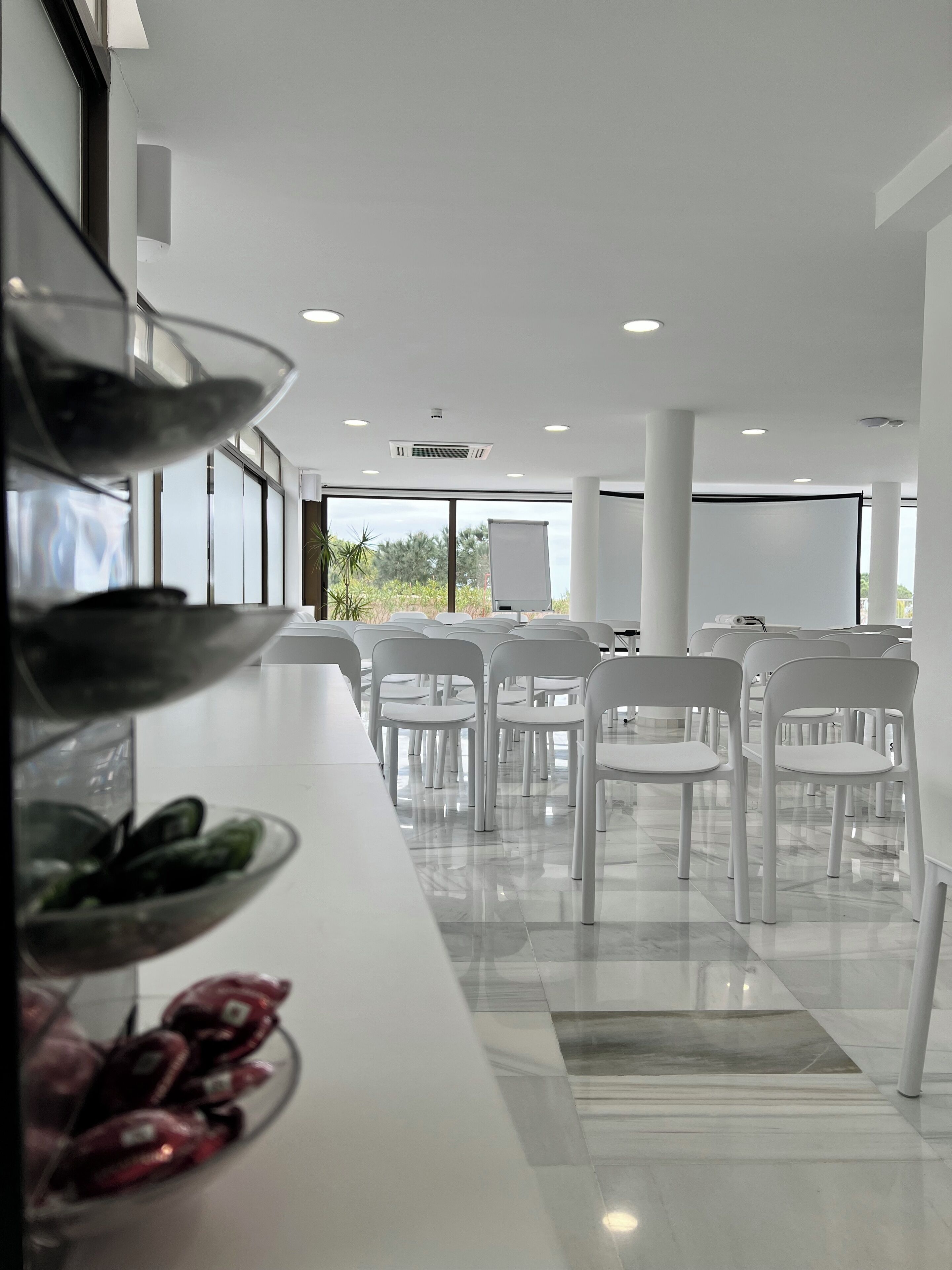 A modern conference room with white chairs and a long table, large windows, and a projector screen.