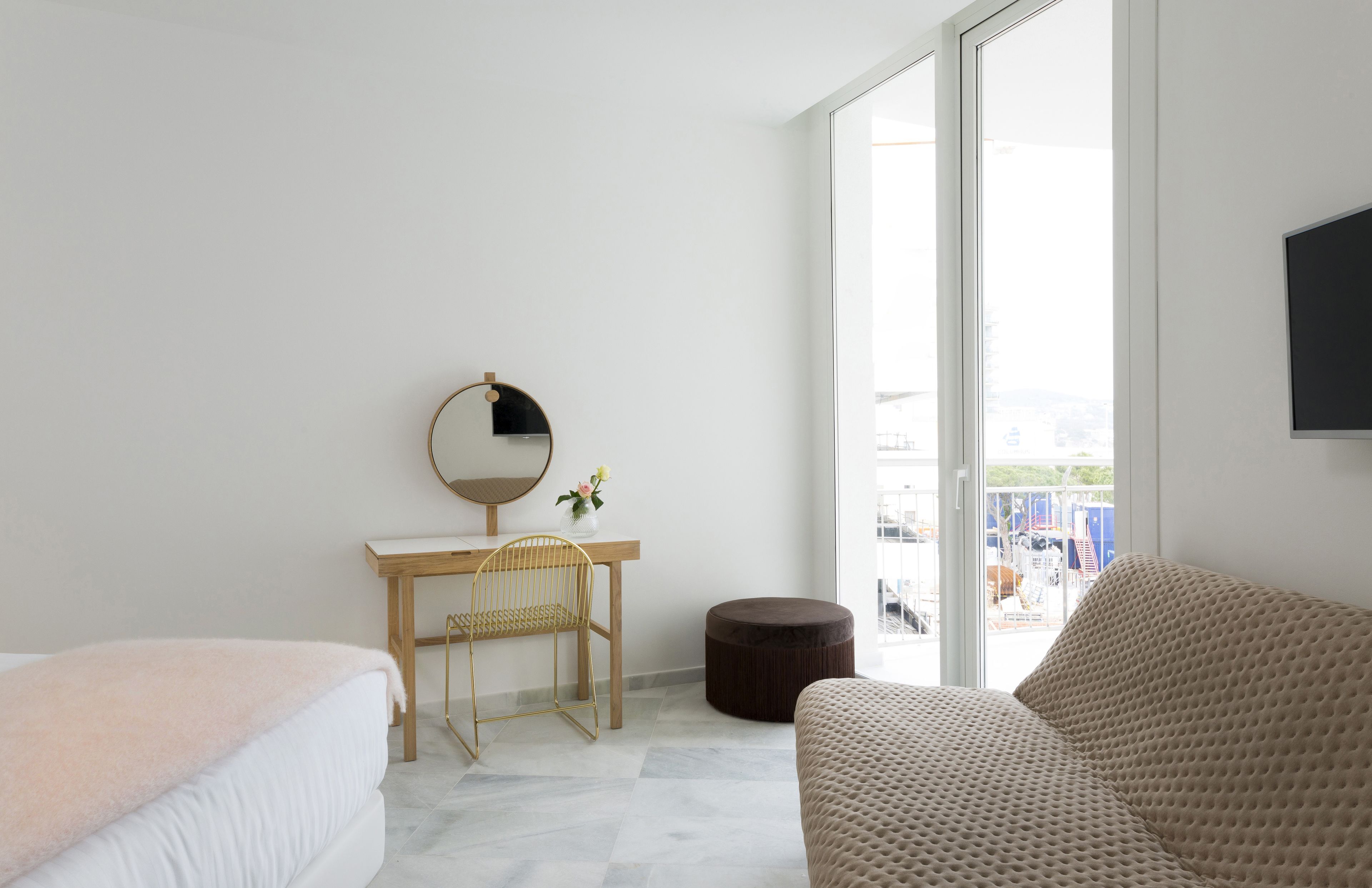 Une chambre d’hôtel moderne comprenant un lit, un bureau avec une chaise, un miroir rond, un pouf rond et une grande fenêtre donnant sur la ville.