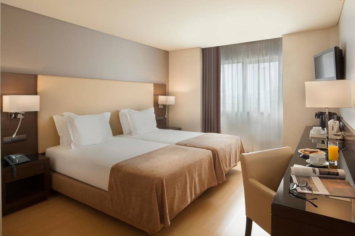 A hotel room with two beds, a desk with a chair, a TV, and a window with curtains.