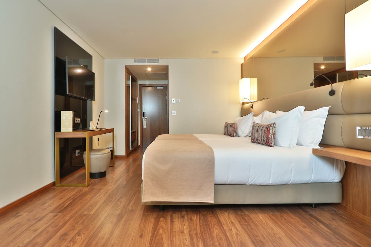 A modern hotel room with a large bed, bedside tables, a TV, and a wooden floor.