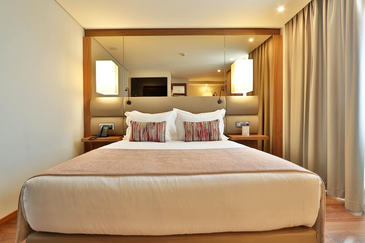 A large bed with a neutral-toned bedspread and two patterned pillows, flanked by nightstands with lamps and a telephone. A mirror reflects the room's interior, and a window with curtains is visible to the right.