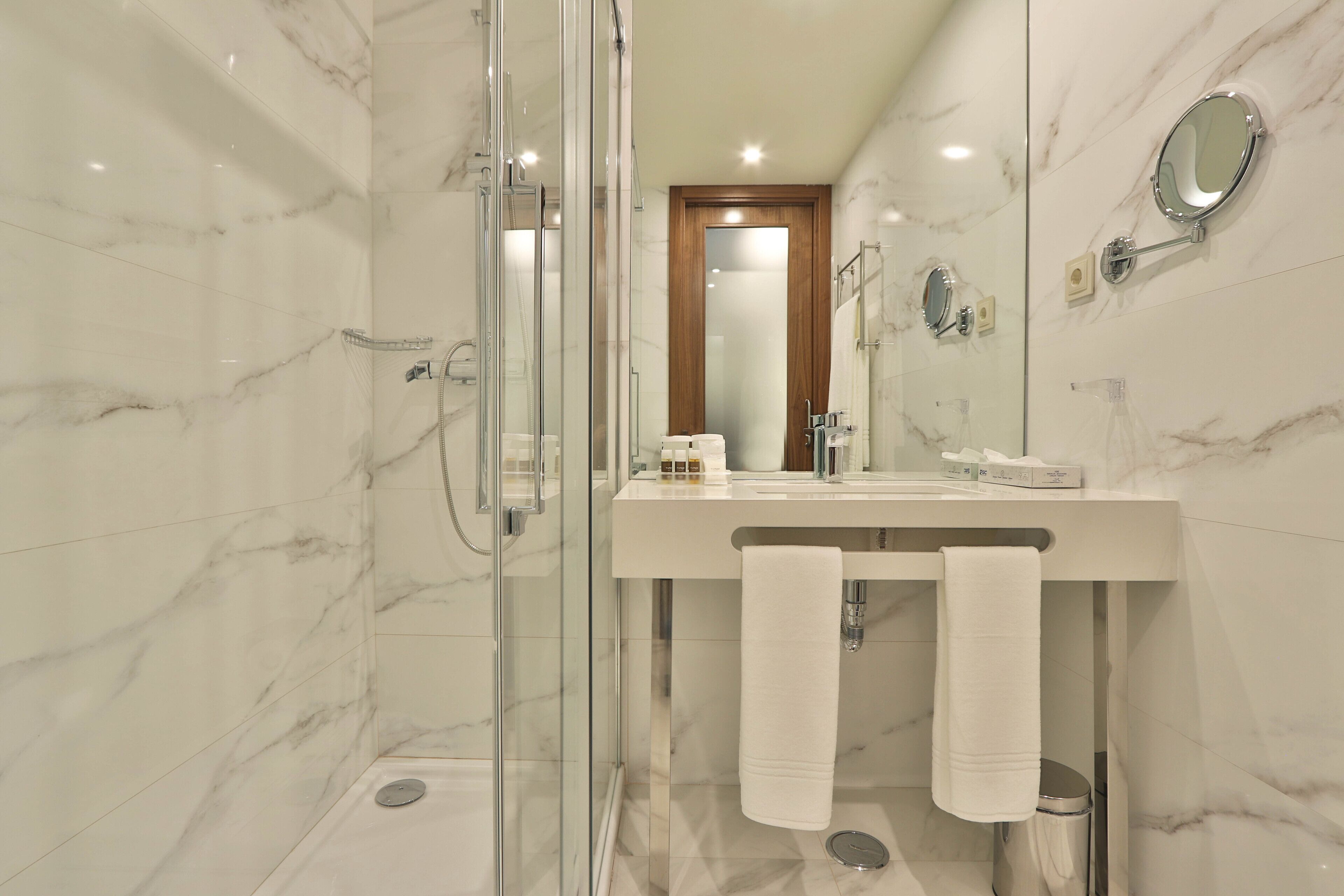 Un baño moderno con cabina de ducha acristalada, pared con azulejos de mármol, puerta de madera y tocador con espejo y perchero.