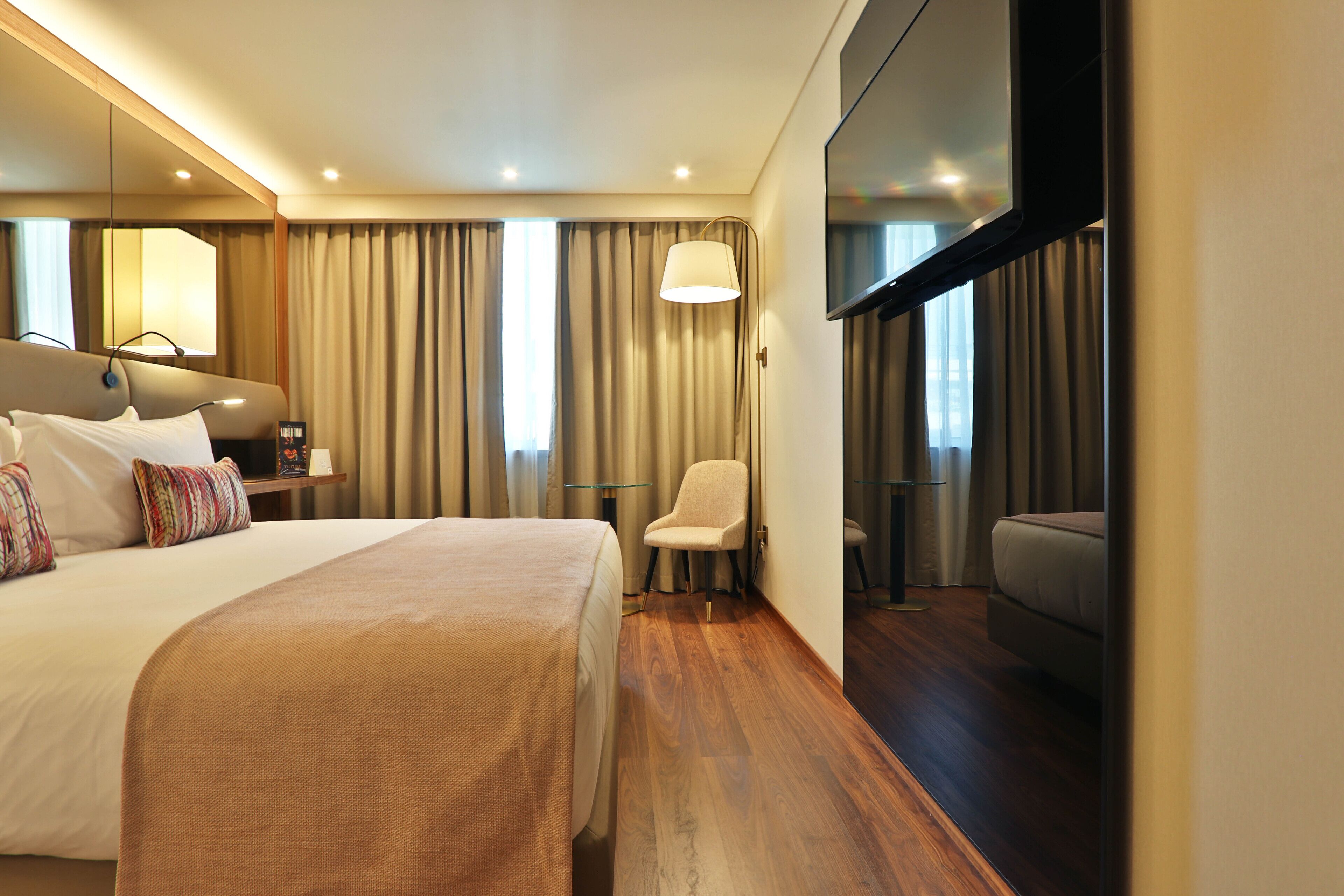 A modern hotel room with a large bed, a seating area, a flat-screen TV, and a large window with curtains.