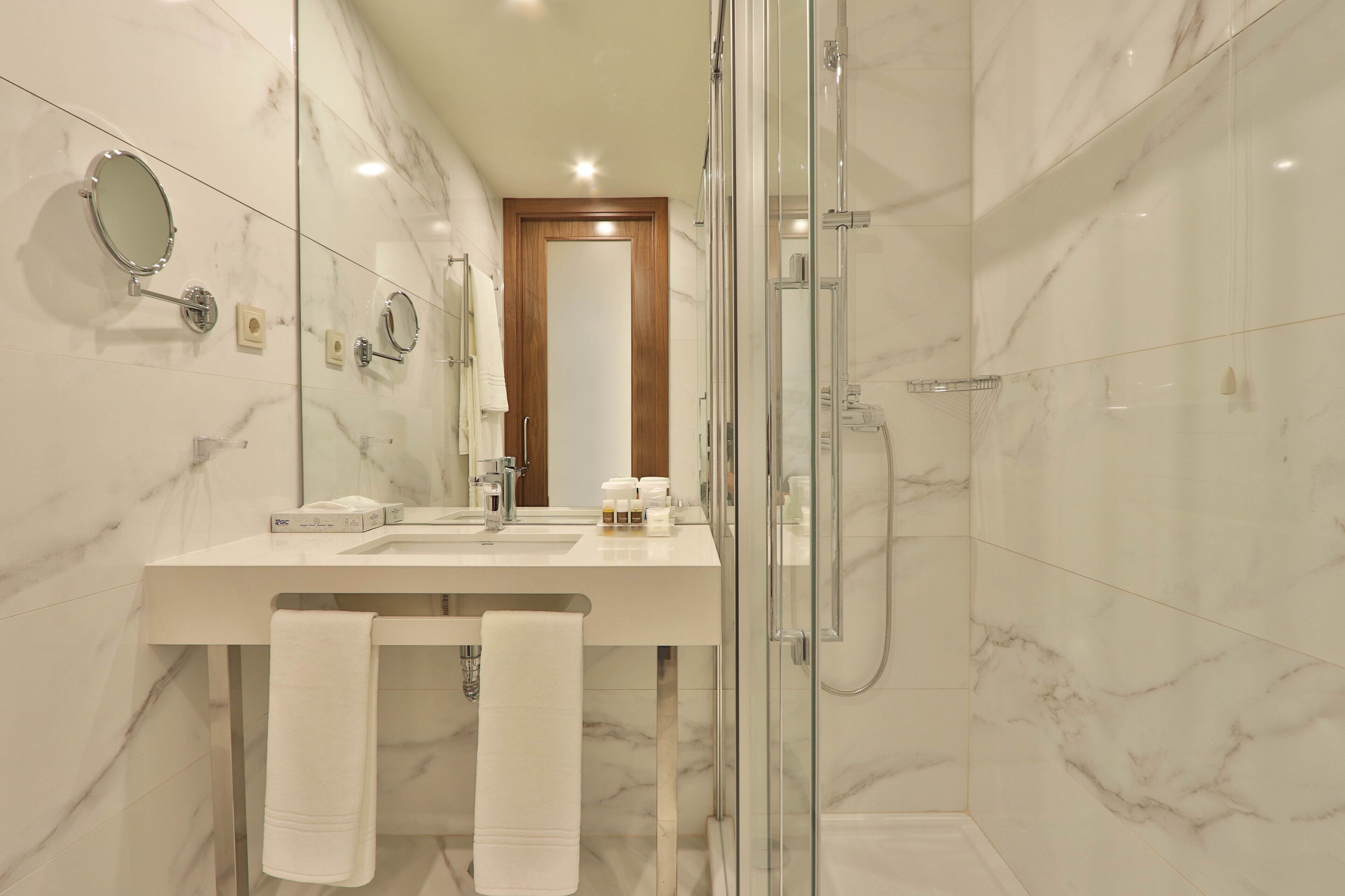 A modern bathroom with a glass shower enclosure, a marble-tiled wall, a wooden door, and a white sink with a mirror and towel rack.