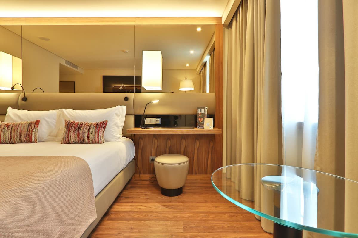 A hotel room with a large bed, a glass table, a small stool, and a wooden desk with a lamp.