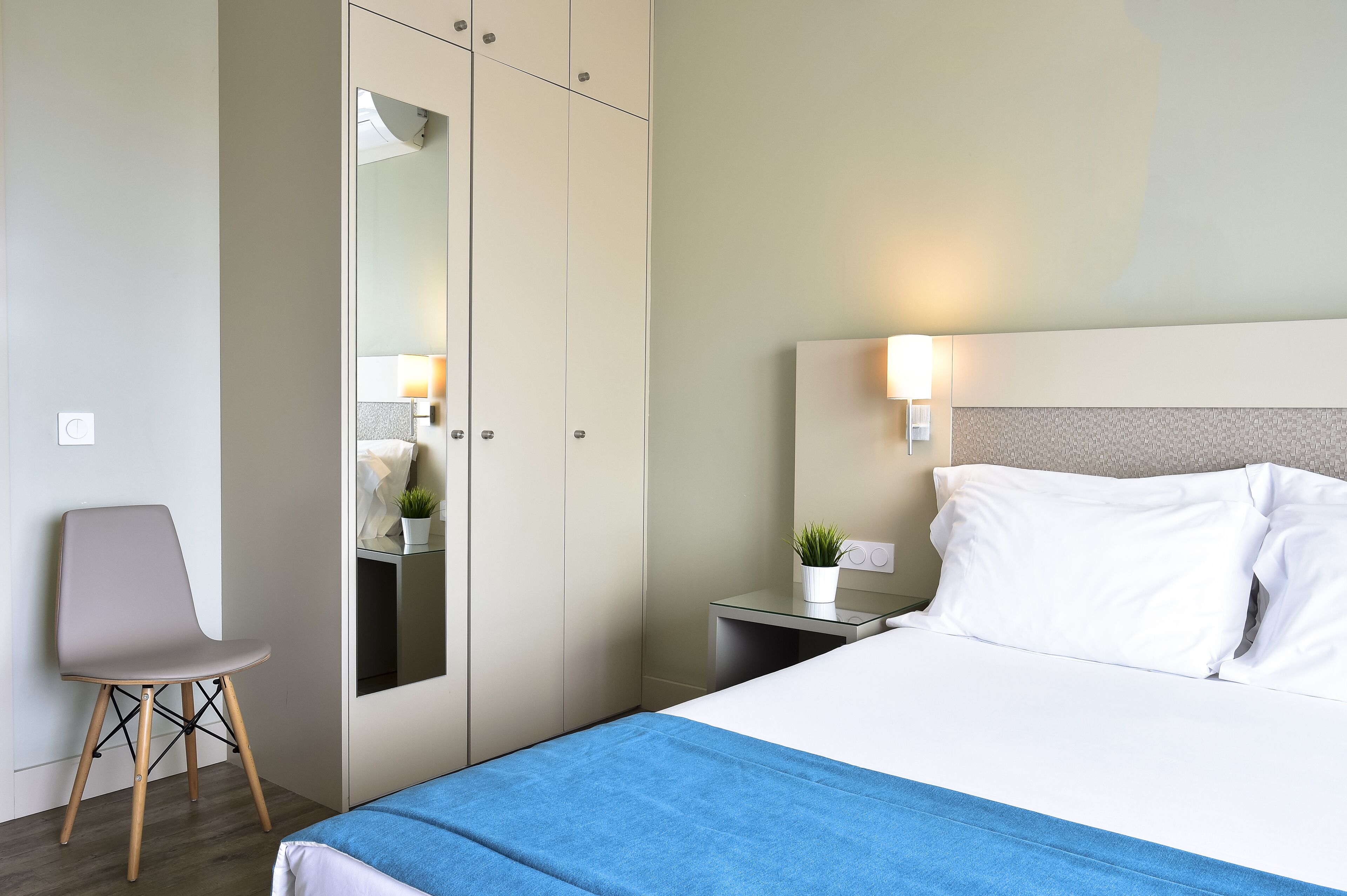A hotel room with a bed, a chair, a wardrobe, a bedside table with a lamp, and a small potted plant.
