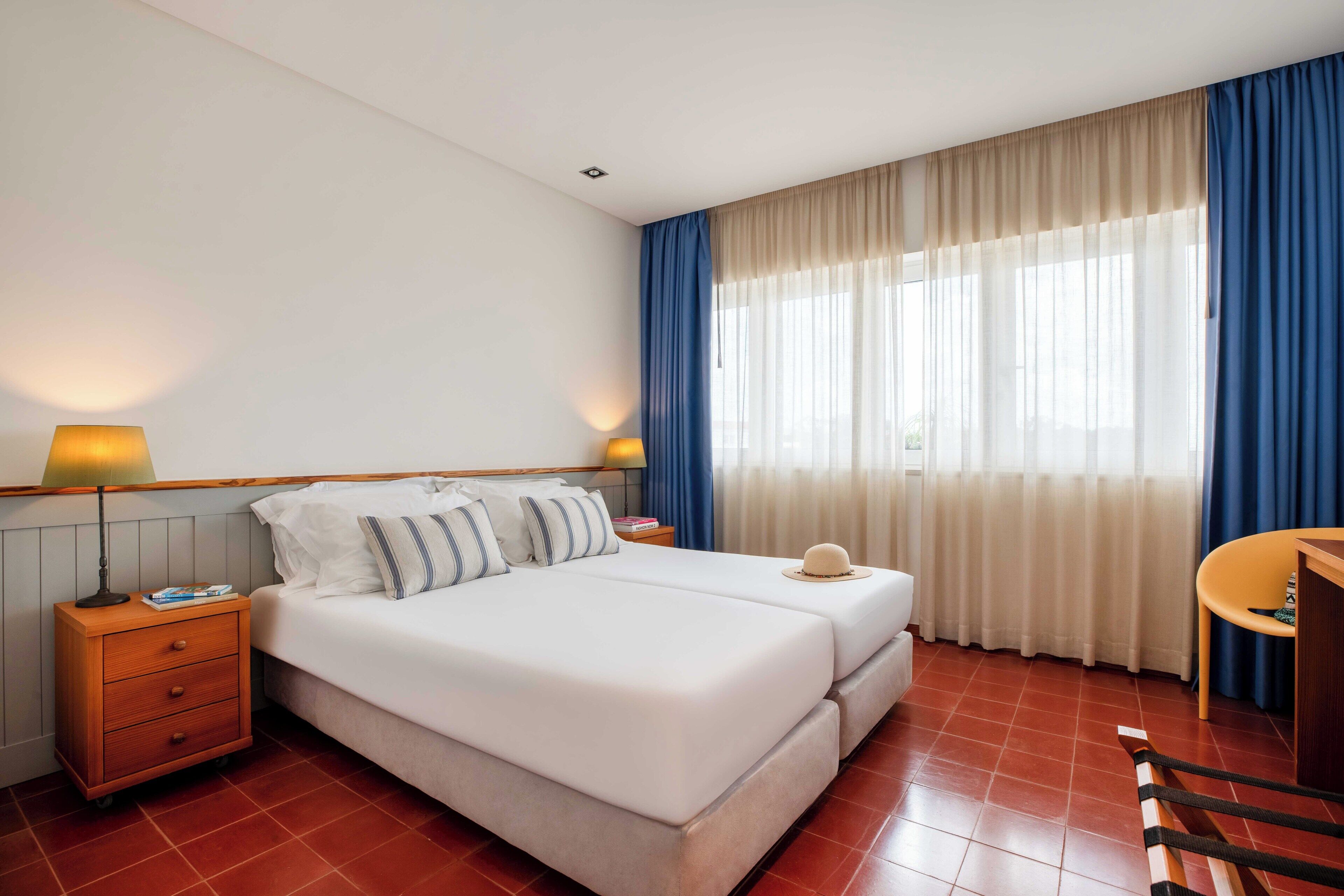A hotel room with a double bed, two bedside tables with lamps, a small desk with a chair, and a large window with curtains.