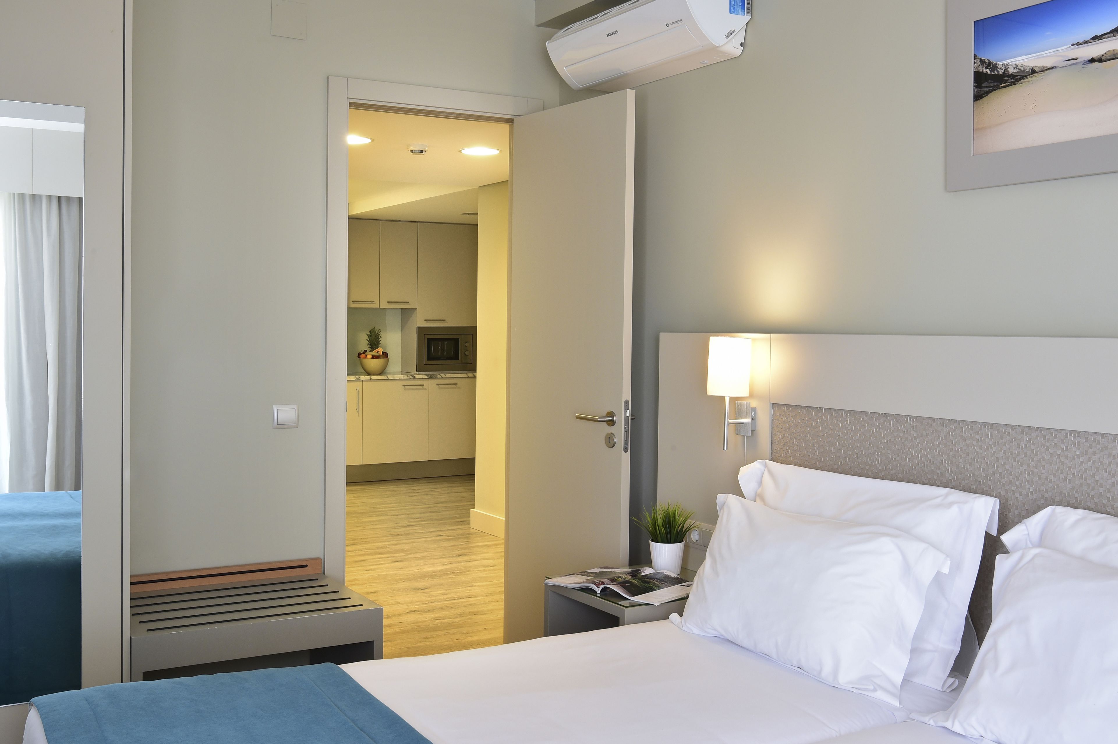 Habitación de hotel con cama, mesita de noche, lámpara y vistas al área de la cocina.