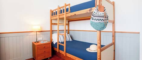 Un lit superposé avec une structure en bois, un matelas bleu et un chapeau blanc sur le lit du bas. On aperçoit une table de chevet en bois avec une lampe, et un sac à motifs accroché au mur.