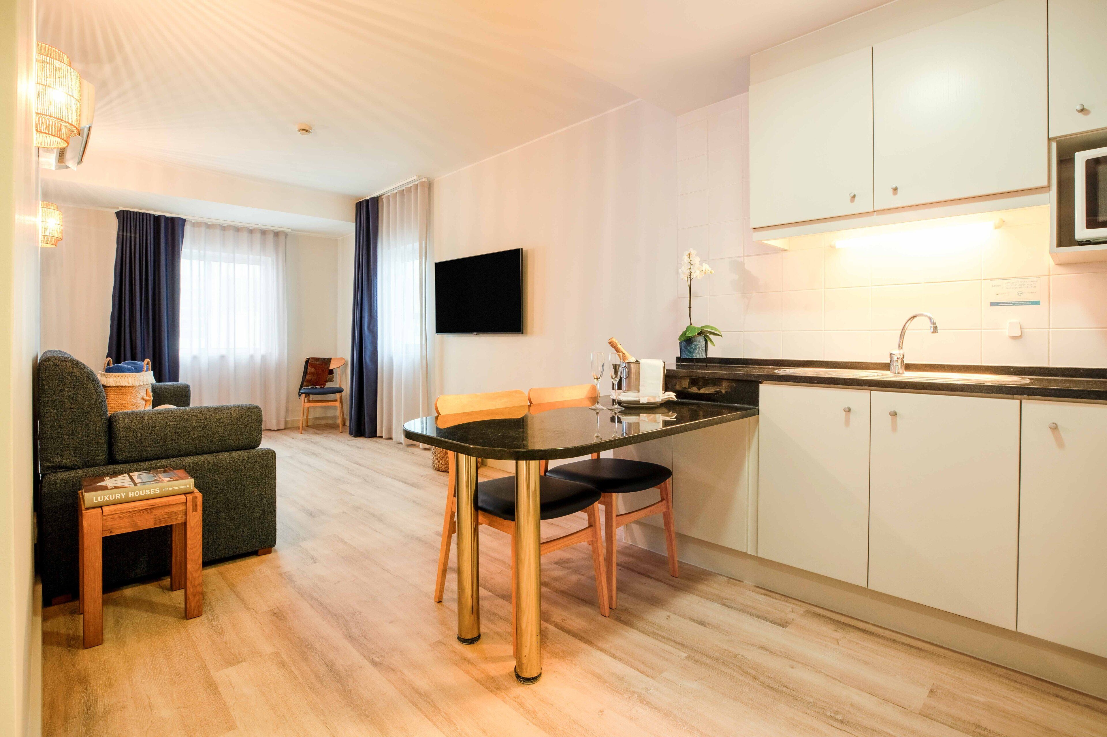 Habitación de hotel moderna con comedor, televisión de pantalla plana, cocina equipada con microondas y un gran ventanal con cortinas.