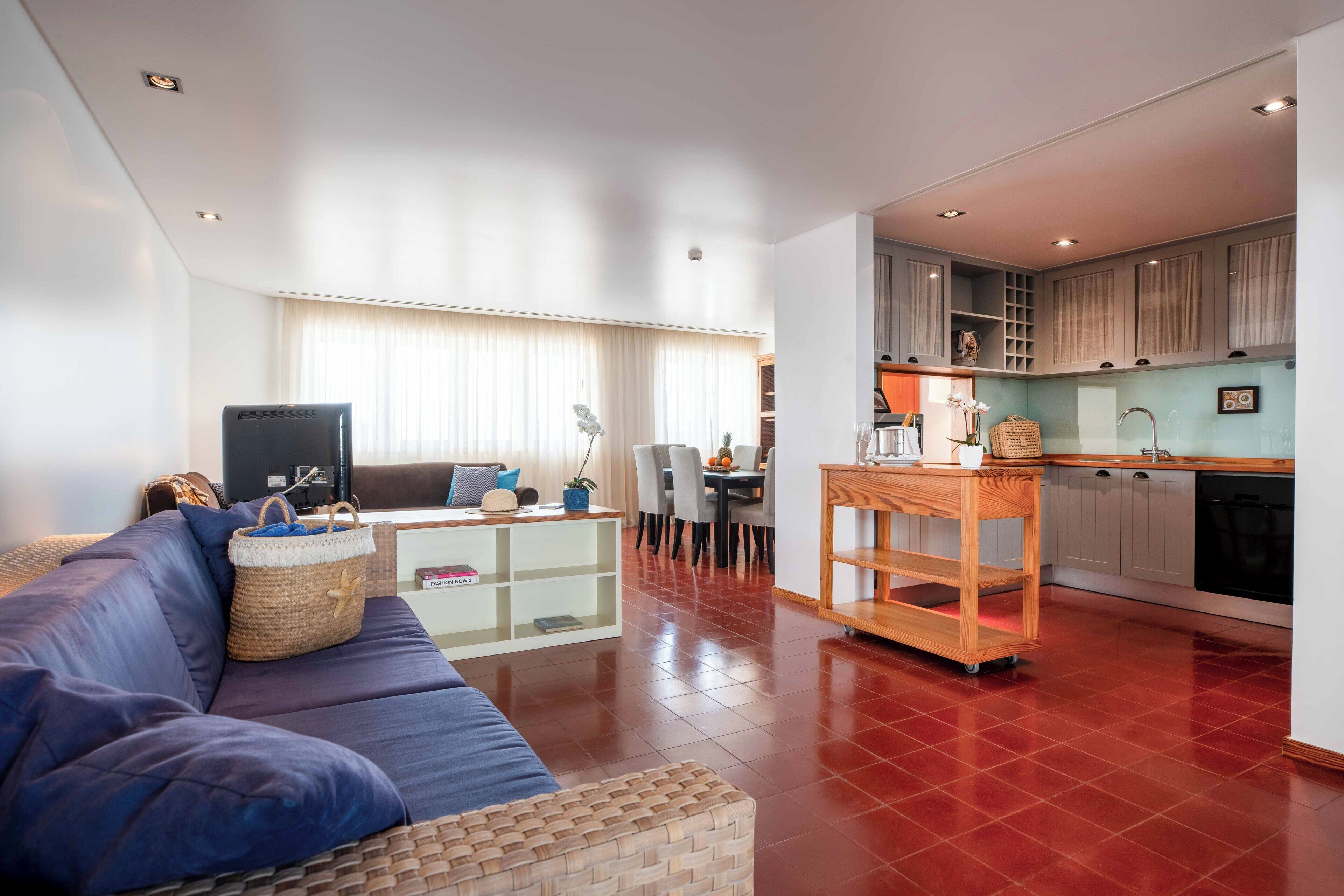 A modern living room with a blue sofa, a wooden coffee table, and a kitchen area with cabinets and a countertop.
