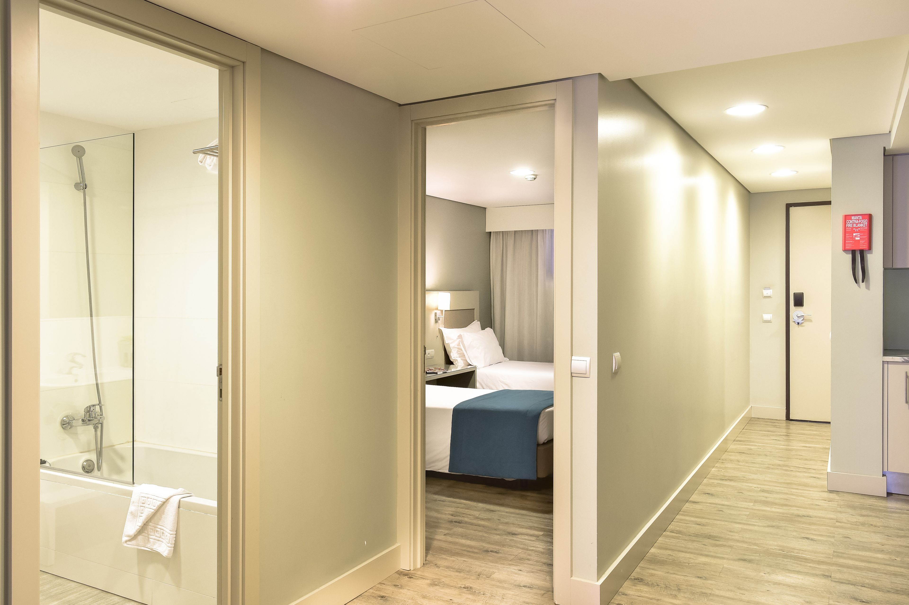 Una habitación de hotel con cabina de ducha de vidrio, cama con manta azul y mesita de noche con lámpara.