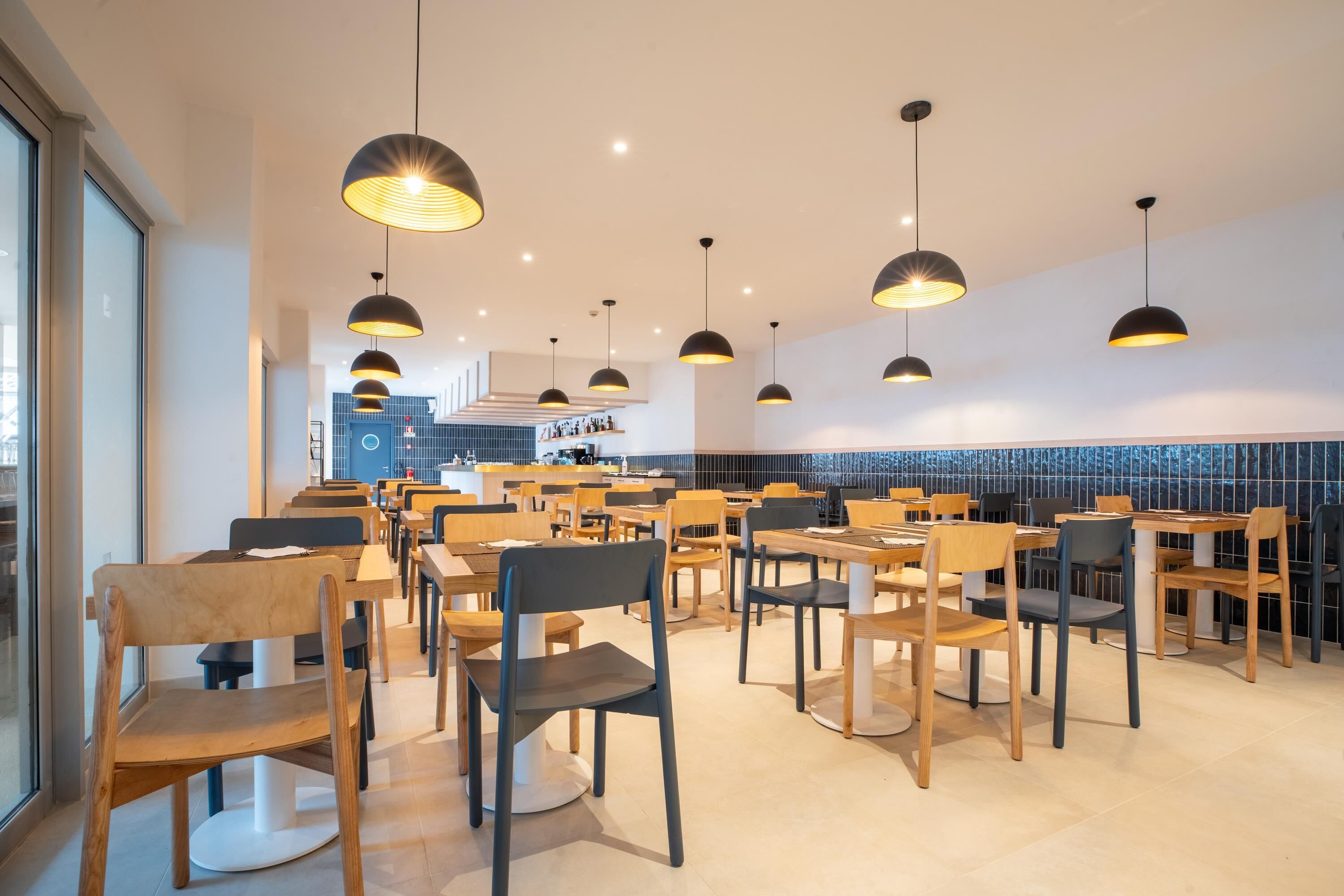 A modern restaurant with multiple tables and chairs, pendant lighting, and a tiled backsplash.