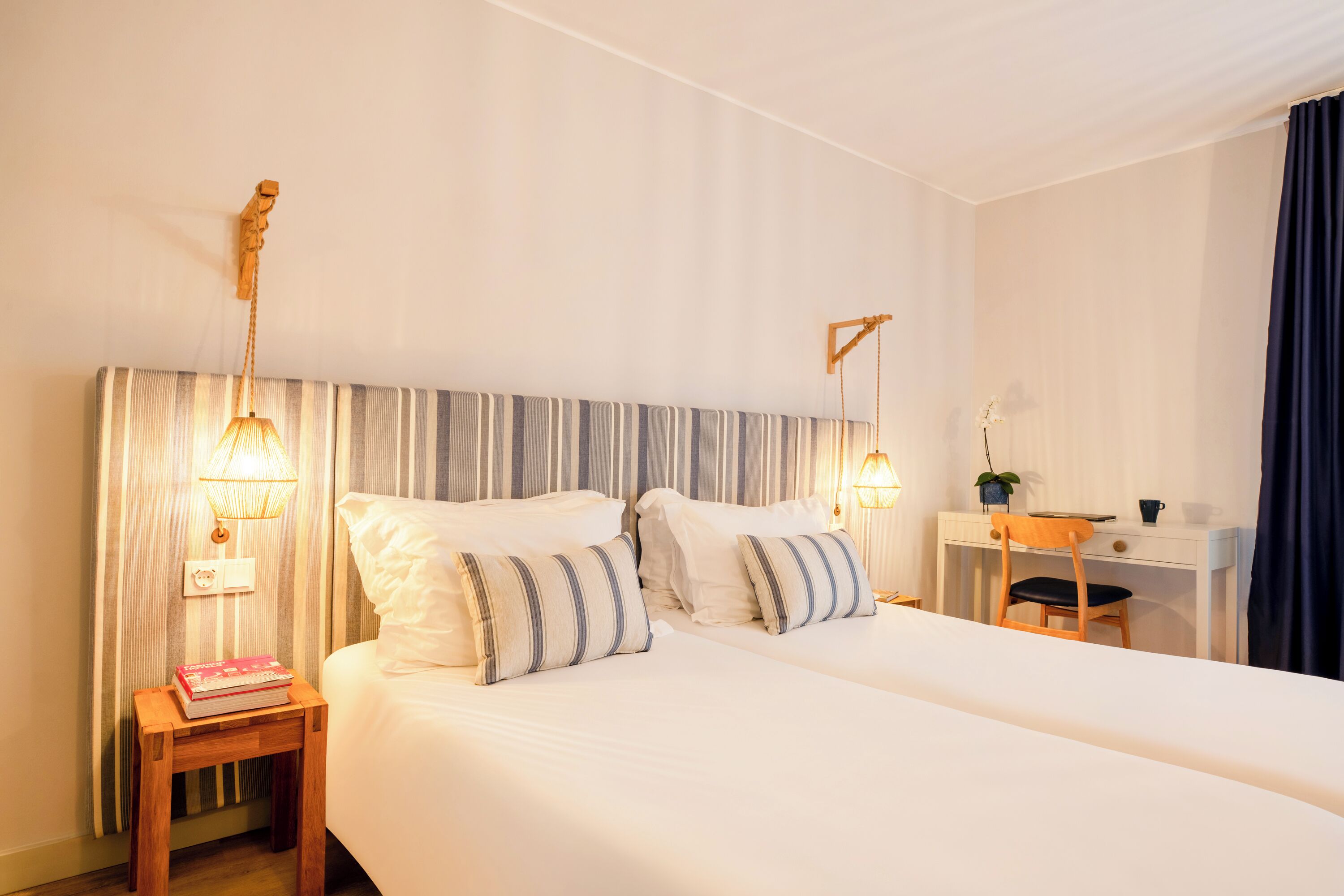 A double bed with striped pillows, a bedside table with a book, a desk with a chair, and two wall-mounted lamps.