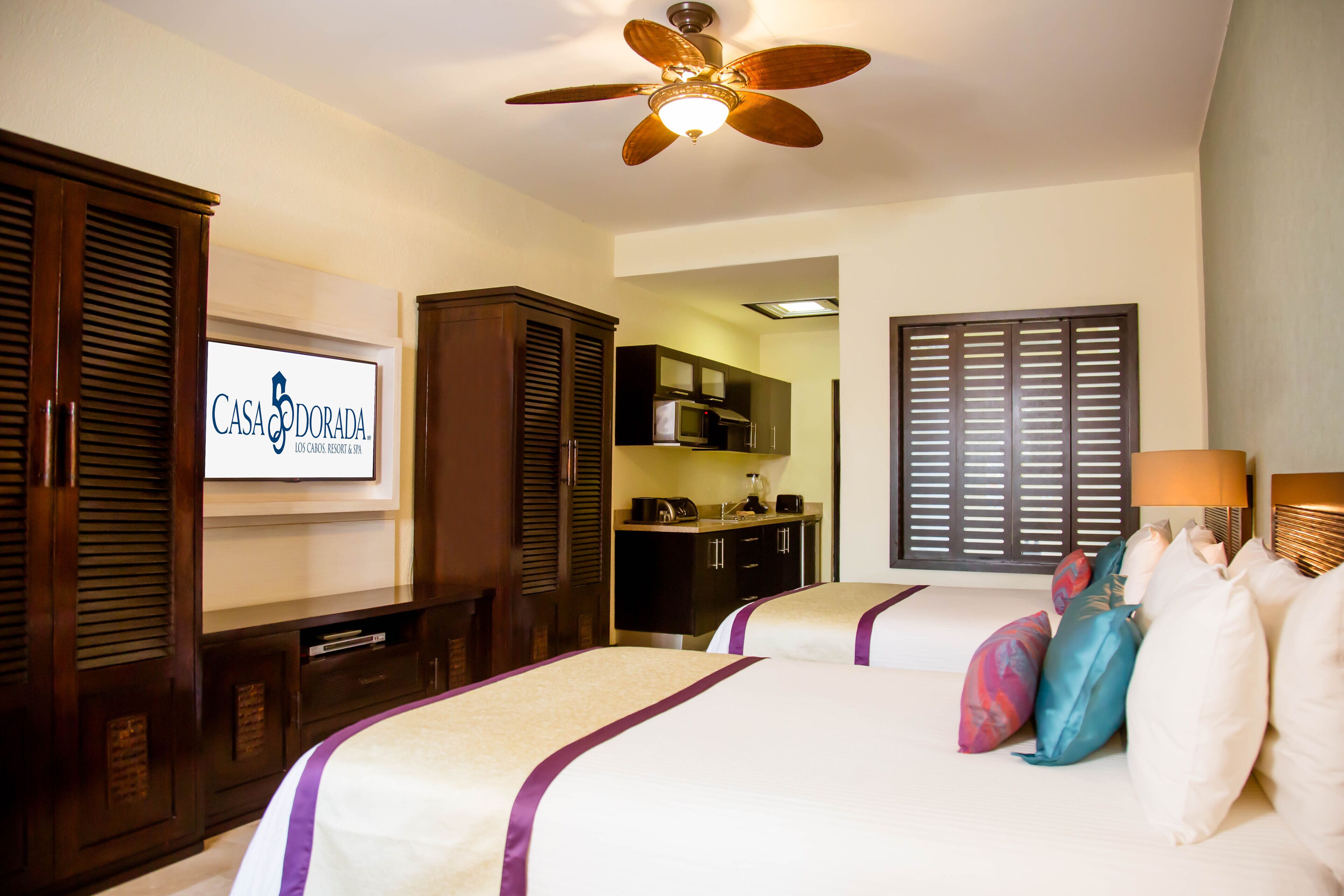 A hotel room with two beds, a ceiling fan, a TV displaying the hotel name, and a kitchenette area.