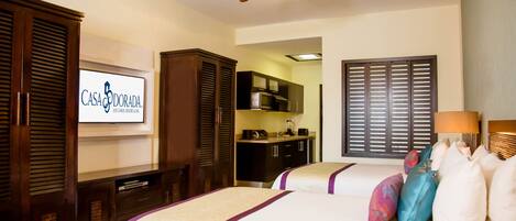 A hotel room with two beds, a ceiling fan, a TV displaying the hotel name, and a kitchenette area.