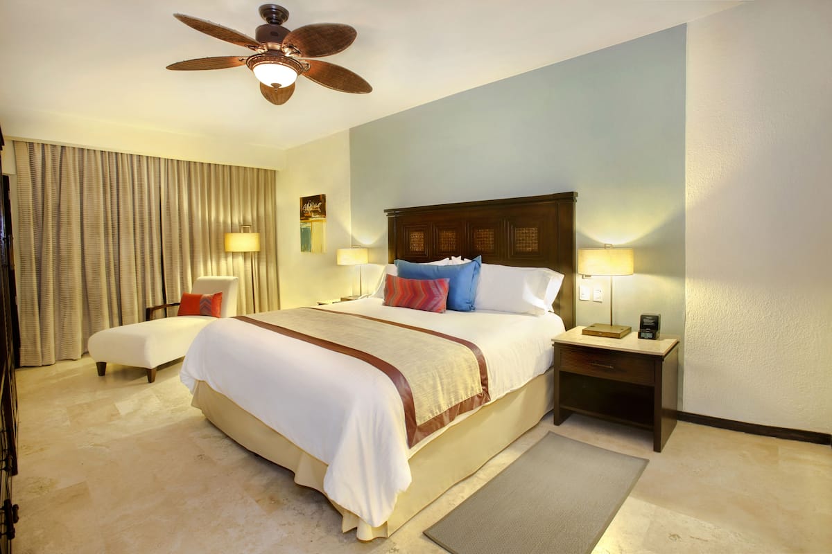 A hotel room with a large bed, a nightstand, a desk, a chair, a ceiling fan, and a view of the city.