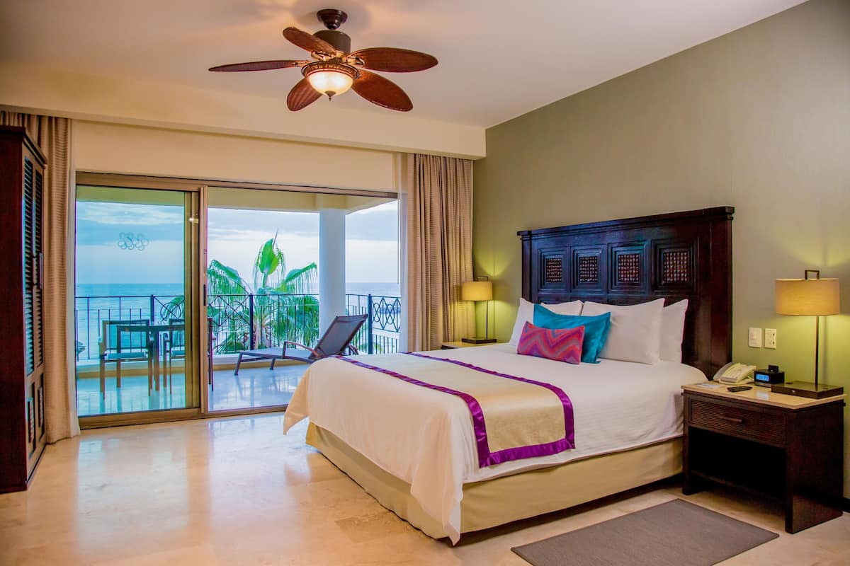 A hotel room with a large bed, a ceiling fan, a nightstand with a lamp, and a view of the outdoors.