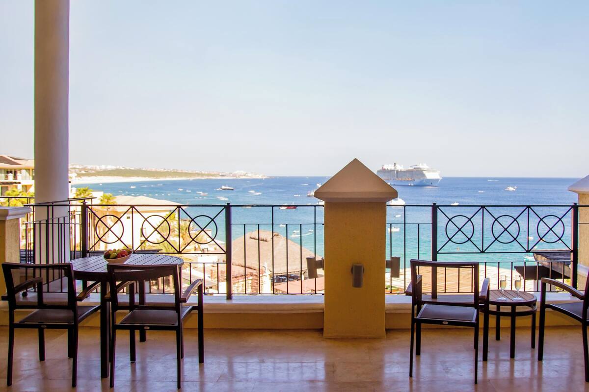 A balcony with a view of the sea, featuring a ship in the distance, a railing, and several chairs and tables.