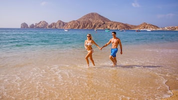 A beach scene with clear water, rocky hills in the background, and boats on the sea.