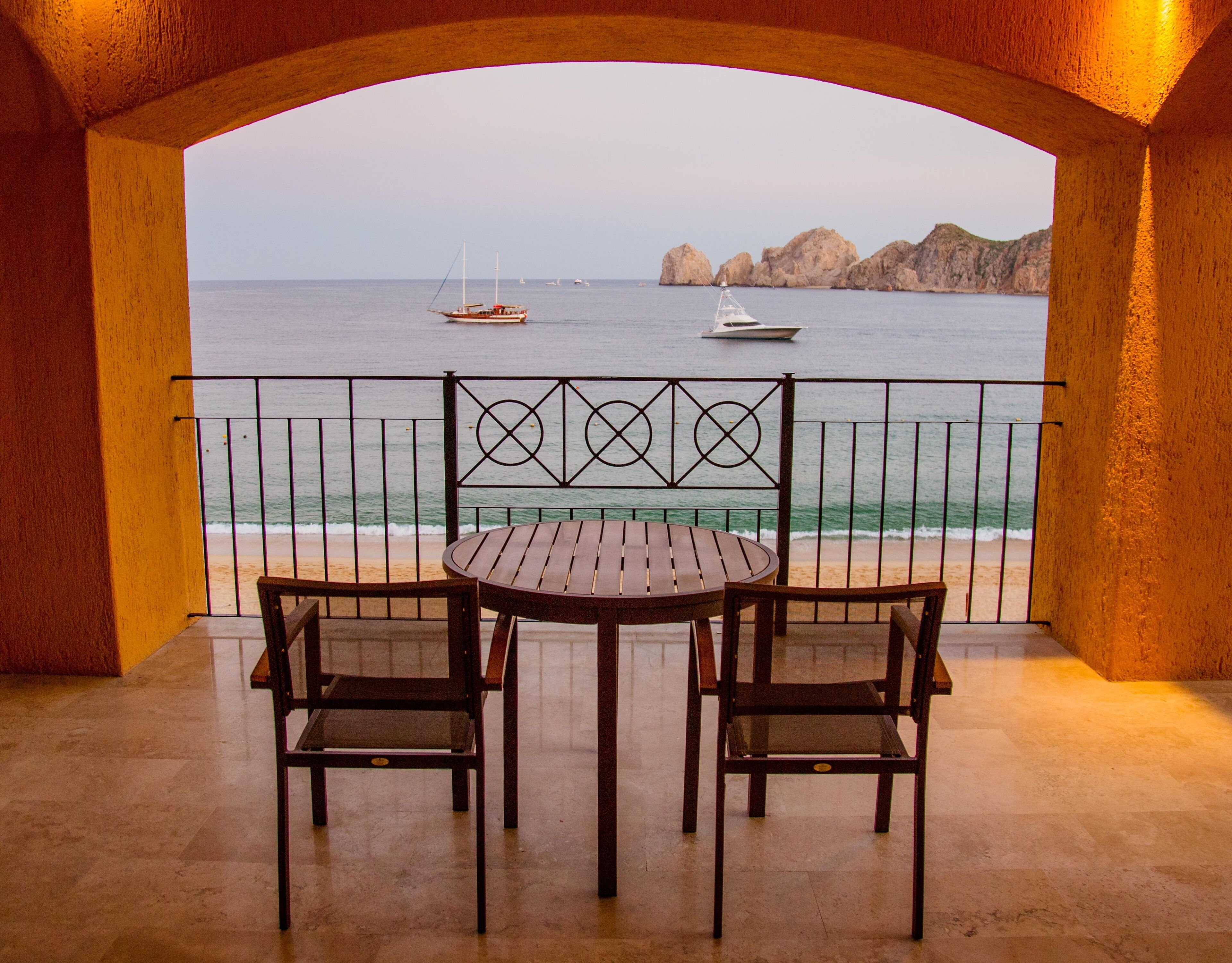 Un balcón con una mesa y dos sillas con vista al mar, mostrando embarcaciones y formaciones rocosas.