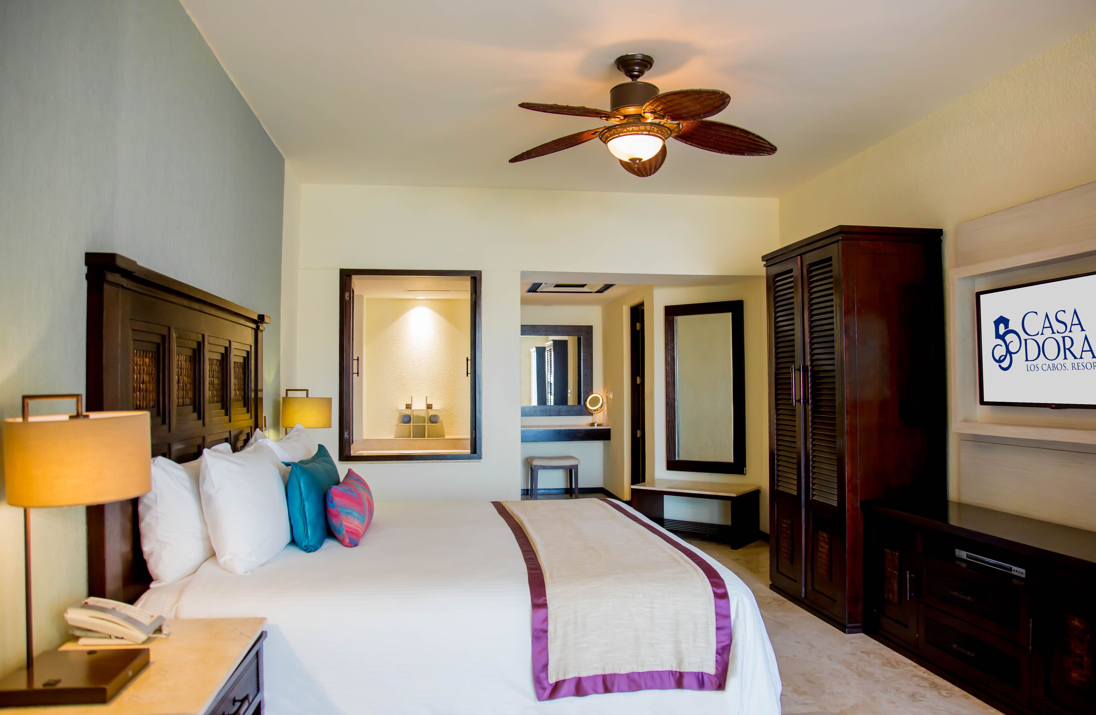 A hotel room with a bed, bedside lamps, a telephone, a television, a ceiling fan, and a mirror.