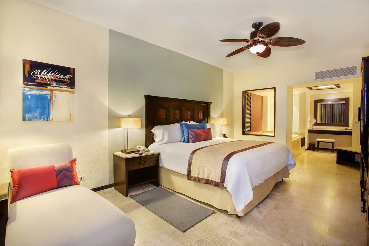 A hotel room with a bed, two bedside tables, a nightstand, a ceiling fan, a mirror, and a painting on the wall.