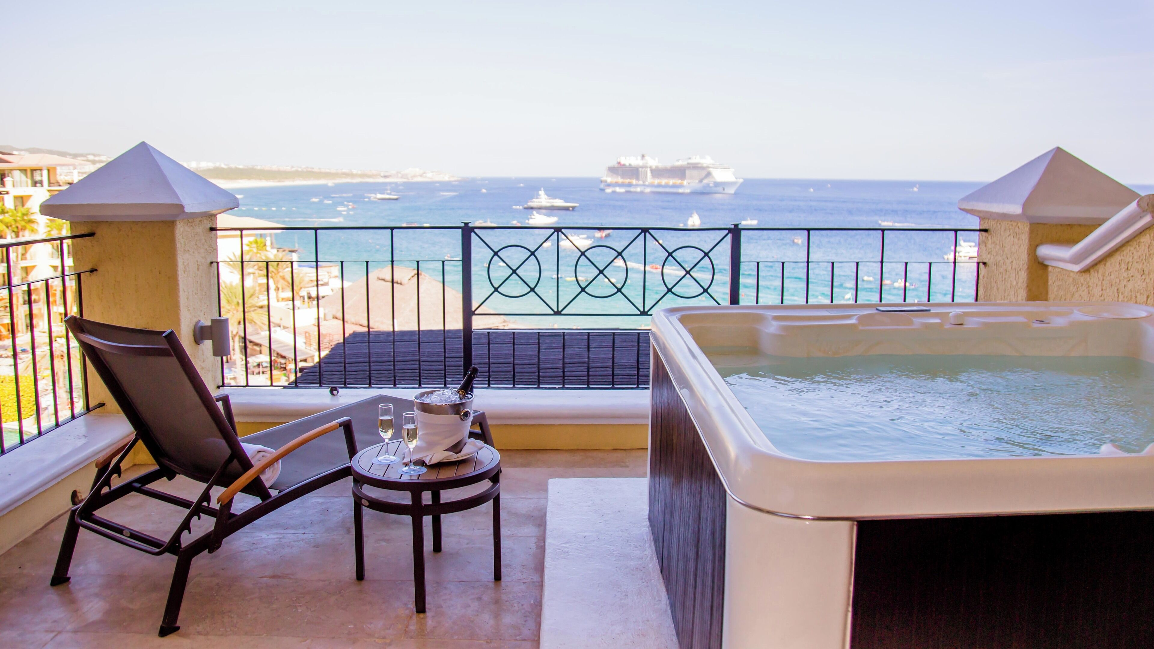 Un balcon doté d’un jacuzzi, d’une chaise et d’une table avec des boissons, offrant une vue sur la mer, avec des bateaux et un navire à l’horizon.