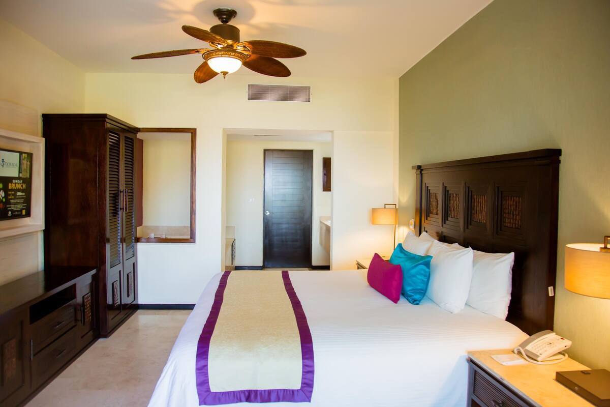 A hotel room with a bed, a desk, a telephone, a nightstand, a ceiling fan, a wardrobe, and a door.