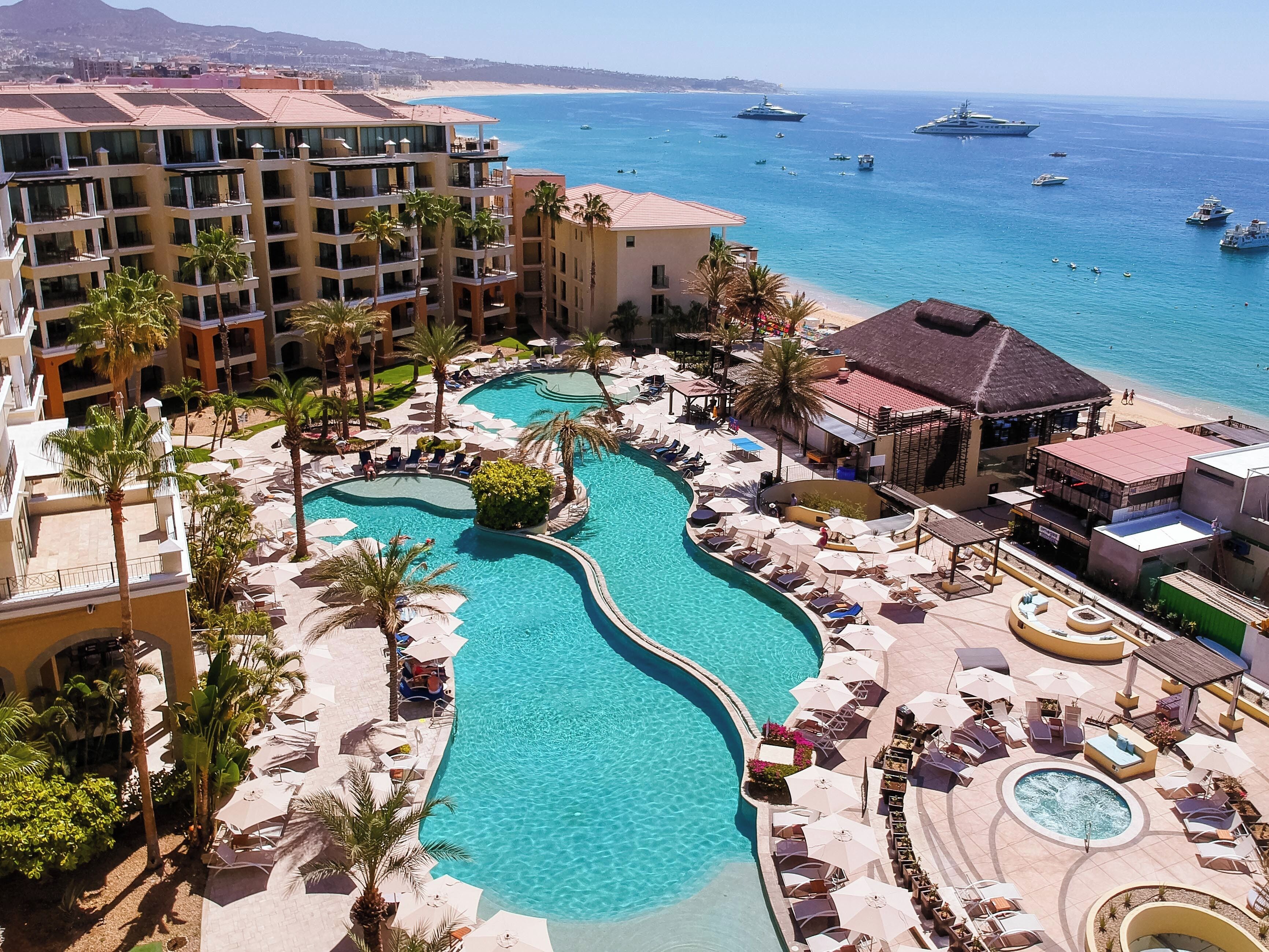 A resort with a central swimming pool, multiple lounge areas with umbrellas, and a view of the ocean with boats.
