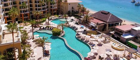 Ein Resort mit einem zentralen Schwimmbad, mehreren Liegebereichen mit Sonnenschirmen und Blick auf das Meer mit Booten.