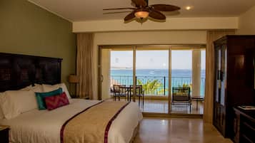A hotel room with a bed, a ceiling fan, a balcony with a view of the ocean, and a desk with a lamp.