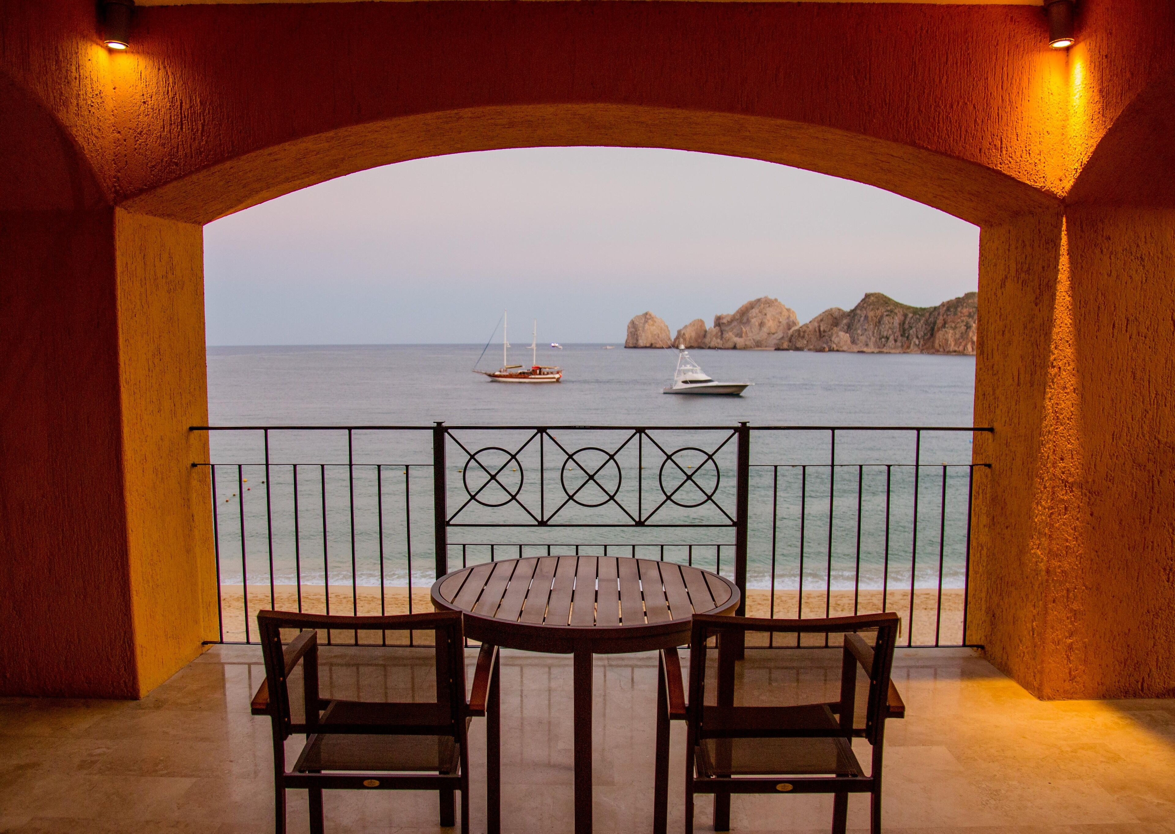 A balcony with a wooden table and two chairs, overlooking a calm sea with boats and rocky cliffs.