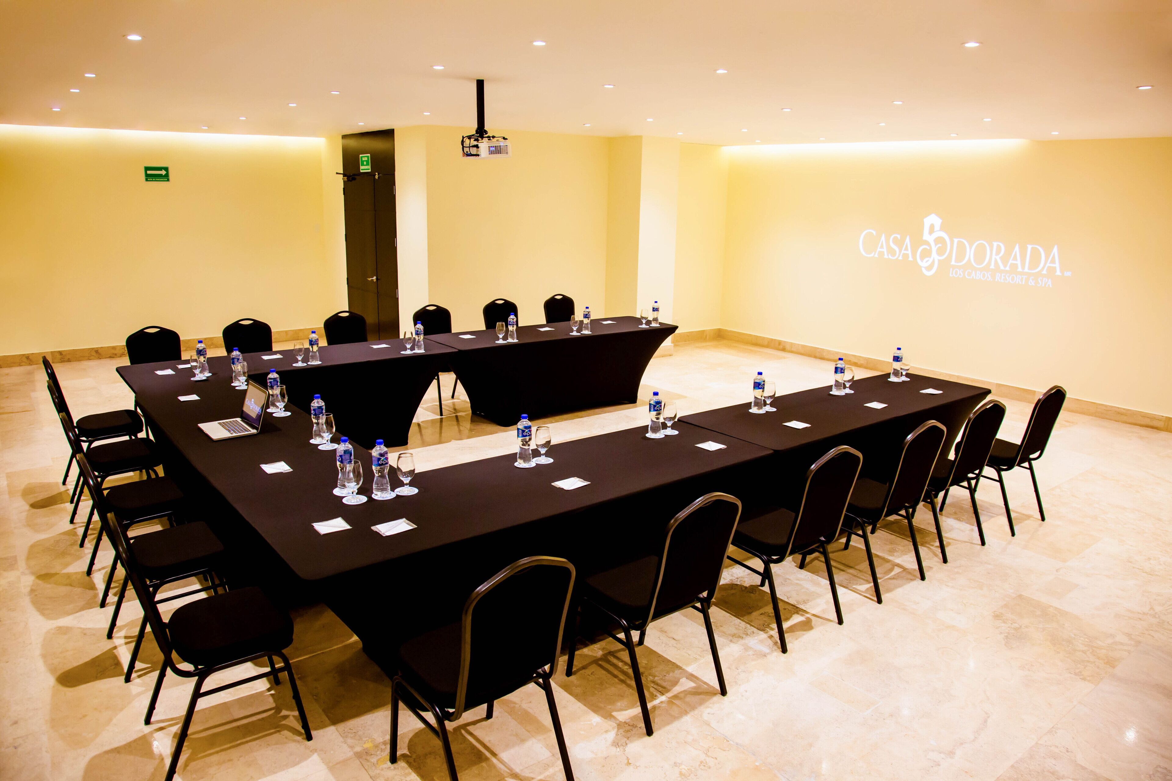 Un salón de conferencias con una larga mesa, sillas, botellas de agua y un ordenador portátil.