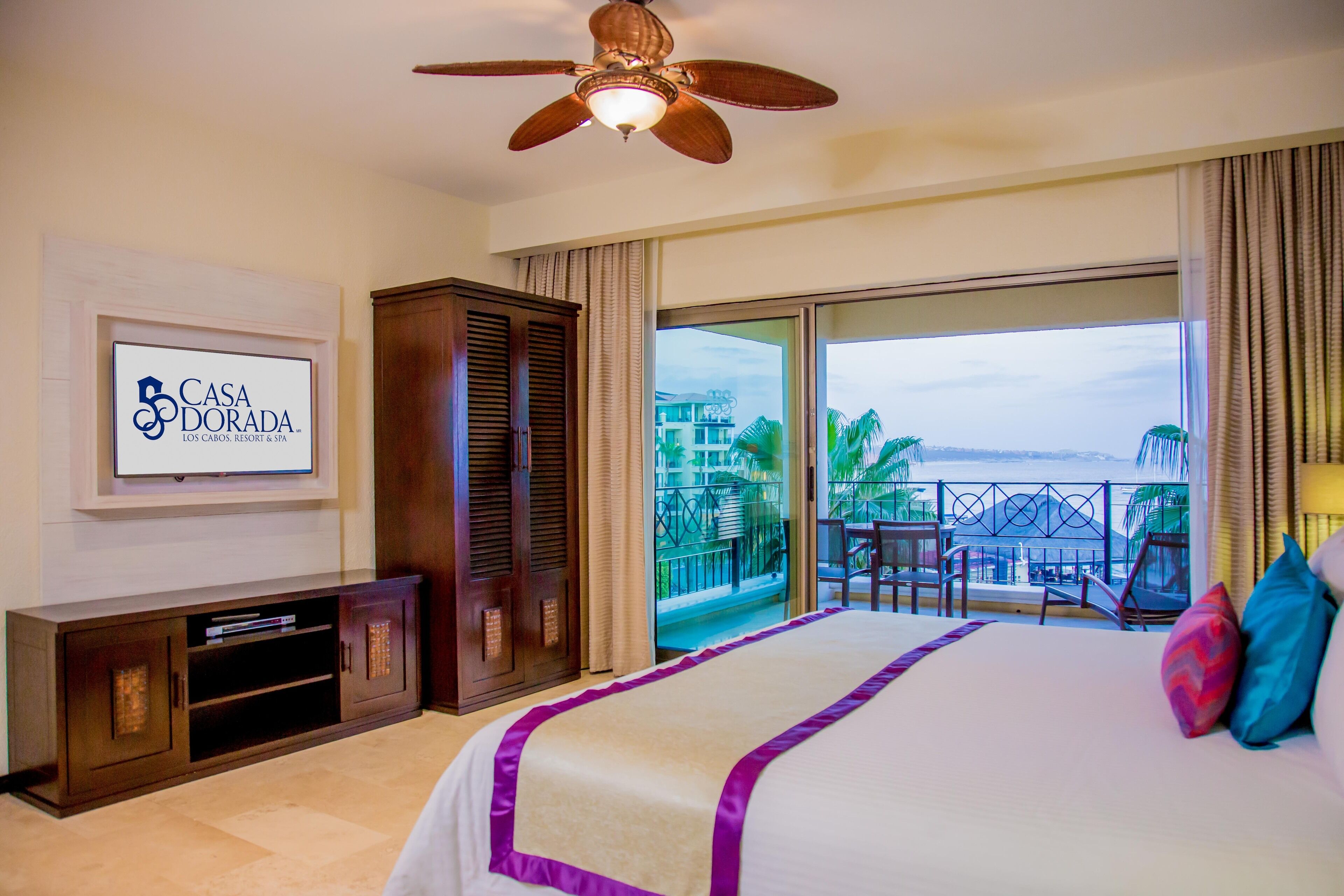 A hotel room with a bed, a television, a minibar, a ceiling fan, and a balcony with a view.