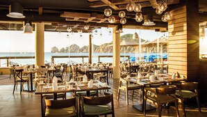 A restaurant with tables set for dining, large windows overlooking the sea, and hanging spherical light fixtures.