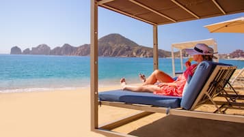 A beach scene with a person relaxing on a sunbed under a shaded area, overlooking the sea and distant mountains.